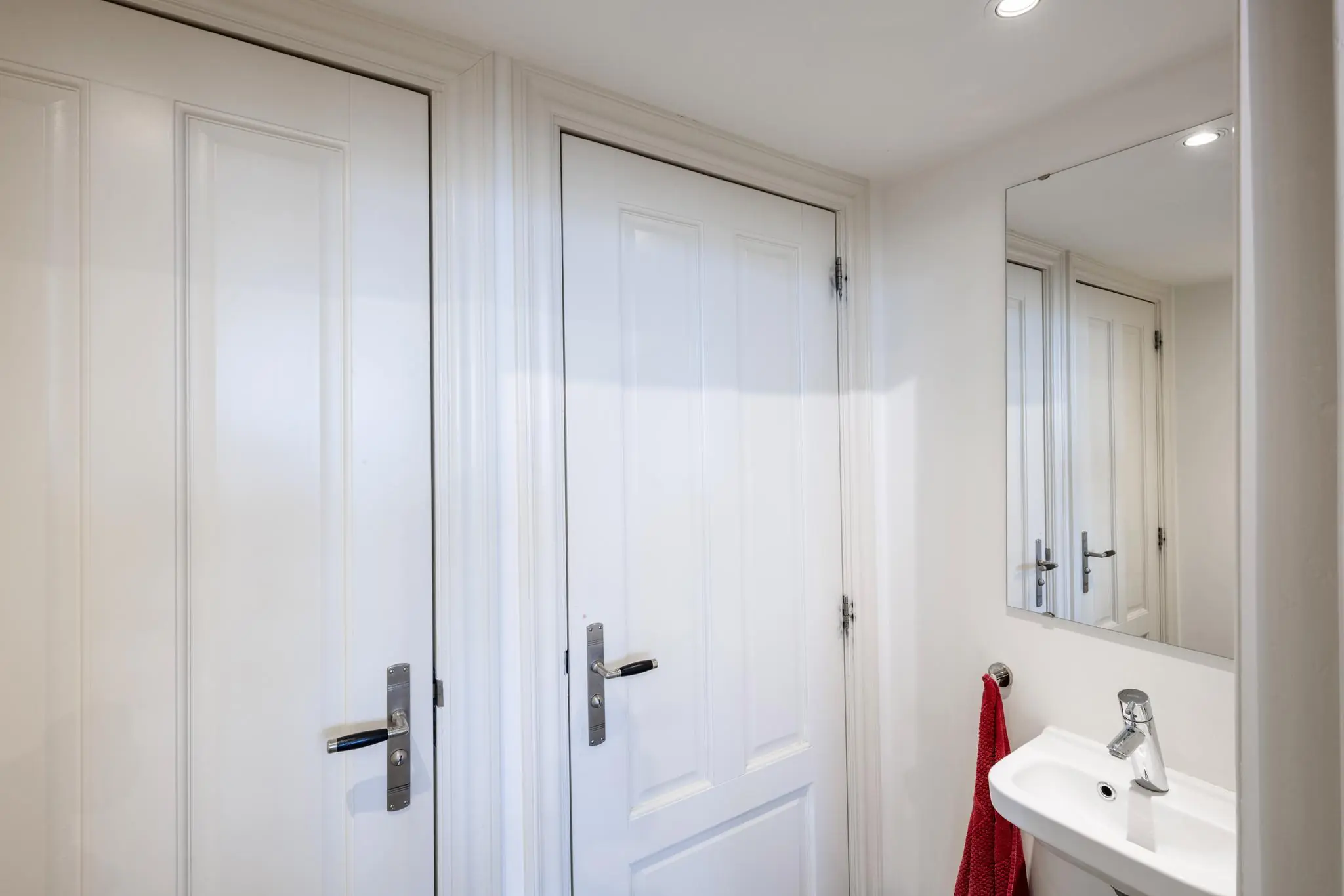 Wit betegelde badkamer met twee witte deuren, een spiegel en een wit fonteintje met rode handdoek.